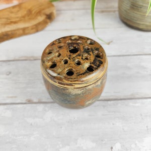 May include: A ceramic flower frog with a speckled, earthy color palette. The round frog has a lid with multiple holes for arranging stems. The exterior features shades of brown, green, and gray. A decorative piece for floral arrangements.