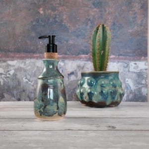 Peut inclure: Un distributeur de savon en céramique avec un vernis brillant moucheté bleu sarcelle et marron, doté d'une pompe en plastique noir et d'un col en liège. À côté se trouve un pot en céramique assorti contenant un cactus vert épineux.