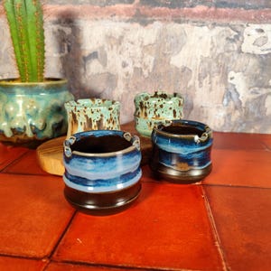 Peut inclure: Ensemble de cendriers en céramique faits à la main de différentes couleurs. Les cendriers ont une forme ronde avec une base marron foncé et un glaçage bleu et turquoise. Un cactus dans un pot turquoise est en arrière-plan.