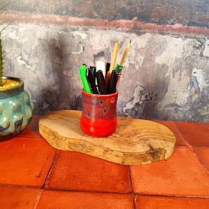 May include: A red and speckled ceramic pen holder filled with pens, pencils, and a small eraser. The holder sits on a wooden slab, with a cactus in a turquoise pot to the left. The background is a textured wall.