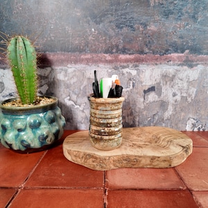 May include: A cylindrical ceramic pencil holder with a ribbed texture and a mottled brown and beige glaze, containing various pens and scissors. It sits on a light-coloured wooden slab next to a green cactus in a blue, textured ceramic pot.