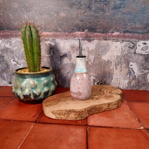 Op de afbeelding: Een keramische olieflacon met een roestvrijstalen tuit, met een roze en blauwe gespikkelde glazuur. Hij staat op een houten serveerplank. Links een groene cactus in een turquoise pot. De achtergrond is een getextureerde muur.