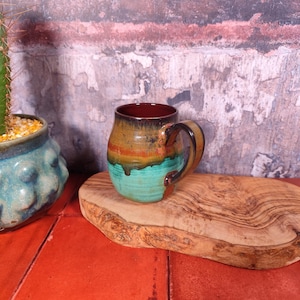 Può includere: Una tazza in ceramica fatta a mano con una smaltatura unica e colorata. La tazza presenta un bordo marrone scuro, che sfuma in tonalità oro, arancione e turchese. Ha un manico robusto e poggia su un tagliere di legno. La tazza è un'opera d'arte funzionale.