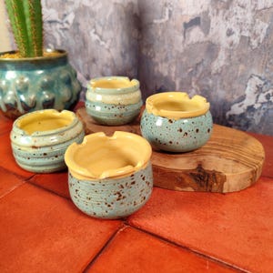 Può includere: Quattro piccoli posacenere in ceramica fatti a mano. Ogni posacenere è rotondo con un interno giallo e un esterno turchese maculato. I posacenere sono esposti su una superficie di legno e una superficie piastrellata rossa. Un cactus in un vaso verde acqua è sullo sfondo.