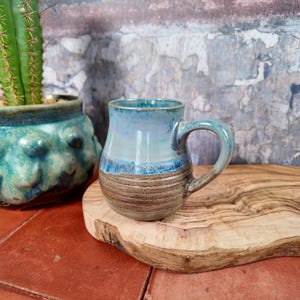 Op de afbeelding: Een handgemaakte keramische mok met een blauw en bruin glazuur. De mok heeft een afgeronde vorm met een oor en staat op een houten oppervlak. Een cactus in een turquoise pot staat op de achtergrond.