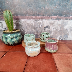 Puede incluir: Un cactus verde en una maceta de cerámica azul verdoso texturizada junto a tres cuencos de cerámica pequeños con esmaltes moteados en tonos azul claro, verde menta y una combinación de rosa y verde, sobre una base de madera.