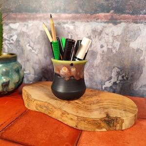 Könnte beinhalten: Ein handgefertigter Stifthalter aus Keramik mit schwarzem Sockel, braun-beigem Mittelteil und grünem Rand. Er enthält Bleistifte, Stifte und Marker. Er steht auf einem ovalen Holzschneidebrett.