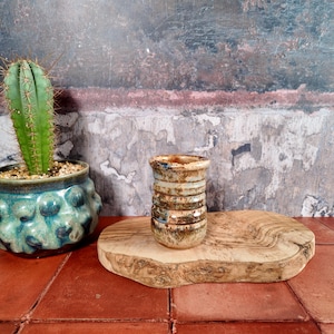 May include: A small, cylindrical ceramic cup with a textured, mottled brown and tan glaze featuring horizontal ridges. The cup has a glossy finish and sits upright, showcasing a rustic appearance with variations in color throughout the surface.