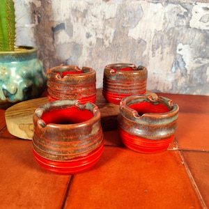 Pode incluir: Quatro cinzeiros de cerâmica feitos à mão. Cada cinzeiro tem um interior e base vermelhos vibrantes, com um exterior texturizado castanho e verde. Os cinzeiros são redondos com uma pequena aba para apoiar os cigarros. Os cinzeiros são exibidos numa superfície castanho-avermelhada.