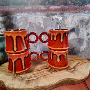 Pode incluir: Quatro canecas de cerâmica empilhadas, em vermelho escuro com esmalte escorrendo laranja e amarelo. Cada caneca tem uma alça circular. As canecas são exibidas em uma superfície de madeira. Altura aproximada: 10 cm.