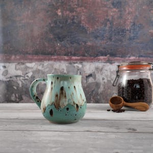 May include: A ceramic mug with a rounded body and a curved handle, featuring a glaze that transitions from a light mint green at the top to a darker teal at the base, accented with vertical brown drips and dark brown speckles throughout the surface.