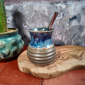 Può includere: Una zucca di yerba mate in ceramica fatta a mano con una glassa blu e marrone. La zucca ha un design a coste e un bordo svasato. Una cannuccia di metallo è inserita nella zucca. La zucca è esposta su un tagliere di legno.