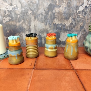 Op de afbeelding: Vier keramische luciferdoosjes met lucifers in verschillende kleuren. De doosjes hebben allemaal een vergelijkbare vorm, met een afgeronde basis en een licht uitlopende bovenkant. Ze zijn versierd met een lichtblauwe glazuur en hebben een subtiel gestreept patroon. De lucifers zijn zwart, rood en blauw.