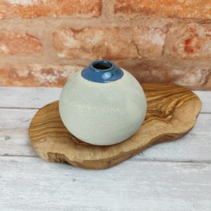 Könnte beinhalten: Eine kleine, runde, weiße Keramikvase mit blauem Rand steht auf einem Holzsockel. Die Vase hat eine gesprenkelte Textur.