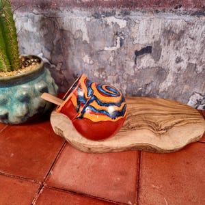 Può includere: Una tazza da mate in ceramica rotonda caratterizzata da uno smalto vivace con motivi vorticosi arancioni, gialli e blu scuro. La tazza presenta un'ampia apertura e una finitura liscia e lucida, con un piccolo cucchiaio di legno all'interno.