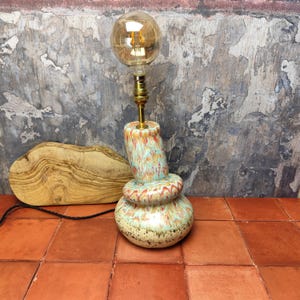 Op de afbeelding: Een keramische tafellamp met een uniek, abstract ontwerp. De lampvoet is een meerkleurige werveling van blauw, groen, oranje en rood. De lampenkap is een heldere glazen bol met een vintage-stijl gloeilamp.