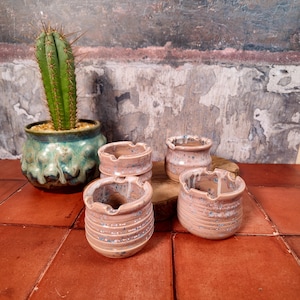 Può includere: Una collezione di posacenere in ceramica fatti a mano in una calda tavolozza di colori terrosi. I posacenere hanno un design strutturato e a coste con sottili accenti blu. Un cactus in vaso aggiunge un tocco di verde.