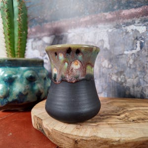 May include: A ceramic cup with a matte black base and a flared rim featuring a reactive glaze in shades of green, brown, and cream. The glaze drips down from the rim, creating a textured, multi-coloured effect against the smooth black body.