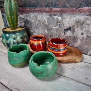May include: Four ceramic bowls with notched rims, including two green bowls with smooth finishes, one orange bowl with a dark drip glaze, and one orange bowl with horizontal blue and dark stripes featuring a small stamped logo on the side.