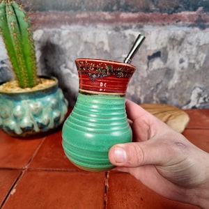 Può includere: Una zucca di yerba mate in ceramica fatta a mano con una cannuccia in acciaio inossidabile. La zucca presenta una combinazione di colori verde, rosso e marrone. La zucca è tenuta in mano, mostrando la sua forma e il suo design unici.