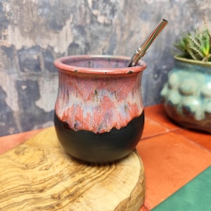 Könnte beinhalten: Eine schwarze und rote Keramik-Mate-Kanne mit einem Edelstahl-Bombilla-Strohhalm. Die Kanne hat eine strukturierte, gesprenkelte Oberfläche.