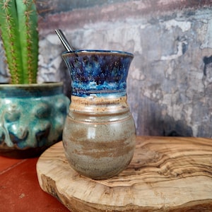 Può includere: Una zucca di yerba mate in ceramica fatta a mano con una glassa blu e marrone. Una cannuccia di metallo è all'interno. La zucca è su una superficie di legno, con un cactus e un vaso turchese sullo sfondo.