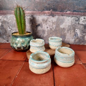 Pode incluir: Quatro cinzeiros de cerâmica pequenos com um esmalte azul claro e creme. Os cinzeiros são redondos com uma superfície texturizada. Um cacto num vaso verde-azulado também está na imagem.