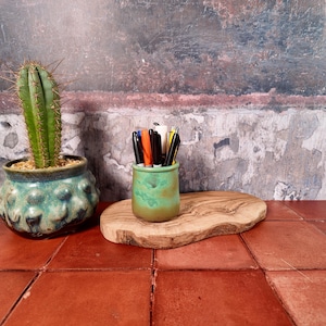 Può includere: Un vaso in ceramica verde contenente un cactus, un portapenne in ceramica verde riempito con varie penne e pennarelli, e una fetta di legno naturale che funge da base per il portapenne, il tutto disposto su una superficie di piastrelle rosse.