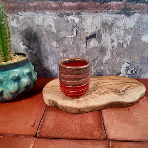May include: A small, handmade ceramic cup with a vibrant red base and a textured, brown upper section. The cup is displayed on a natural wood slab, with a turquoise ceramic pot and a cactus in the background. The cup is approximately 7.6 cm tall.