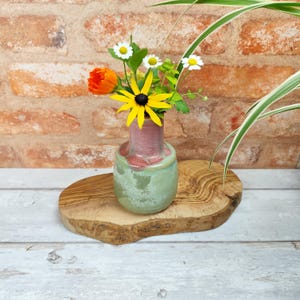 Può includere: Un piccolo vaso in ceramica verde e rosa con una base in legno rustica, riempito con un bouquet di fiori selvatici gialli e arancioni.