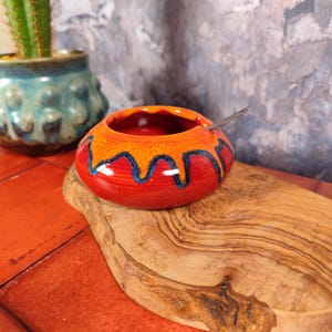Peut inclure: Un petit bol rond en céramique rouge avec des accents décoratifs orange et bleus. Une petite cuillère en argent repose à l'intérieur du bol. Le bol est posé sur une planche à découper en bois. Un pot en céramique turquoise avec un cactus est en arrière-plan.