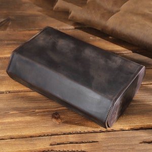 May include: A dark brown leather rectangular box with rounded edges. The box is made of a soft, supple leather and has a smooth, polished finish.