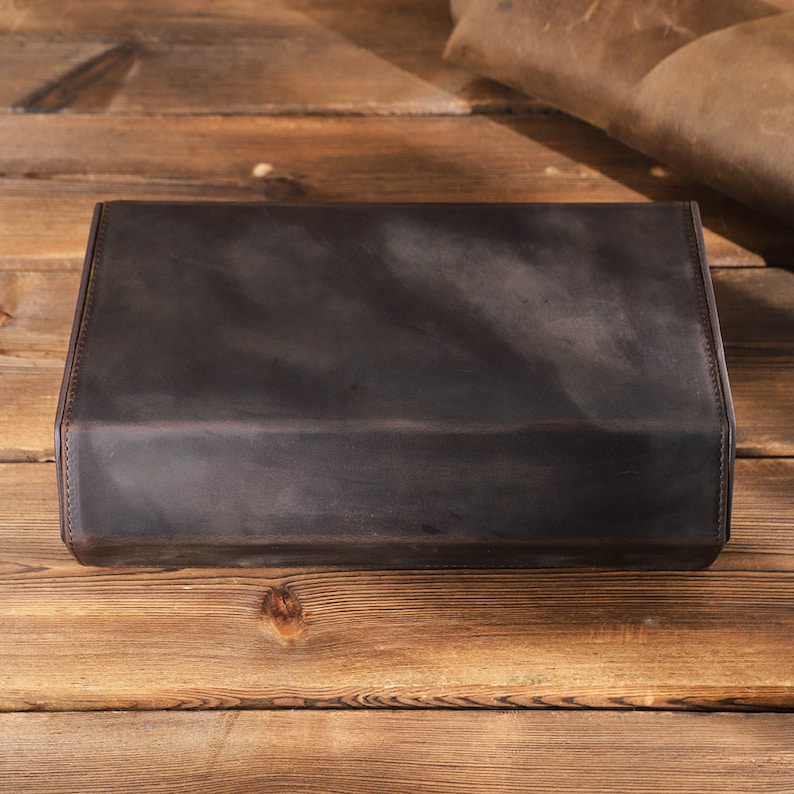 May include: A dark brown leather box with a smooth, textured surface. The box is closed and sitting on a wooden surface.