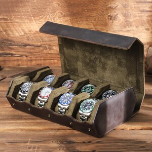 May include: A brown leather watch box with a green velvet interior. The box is open and has eight slots for watches. There are seven watches in the slots, each with a different colored face.