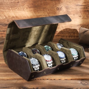 May include: A brown leather watch box with a green velvet interior. The box has six slots for watches and is open to reveal the watches inside. The watches are all different styles and colors.