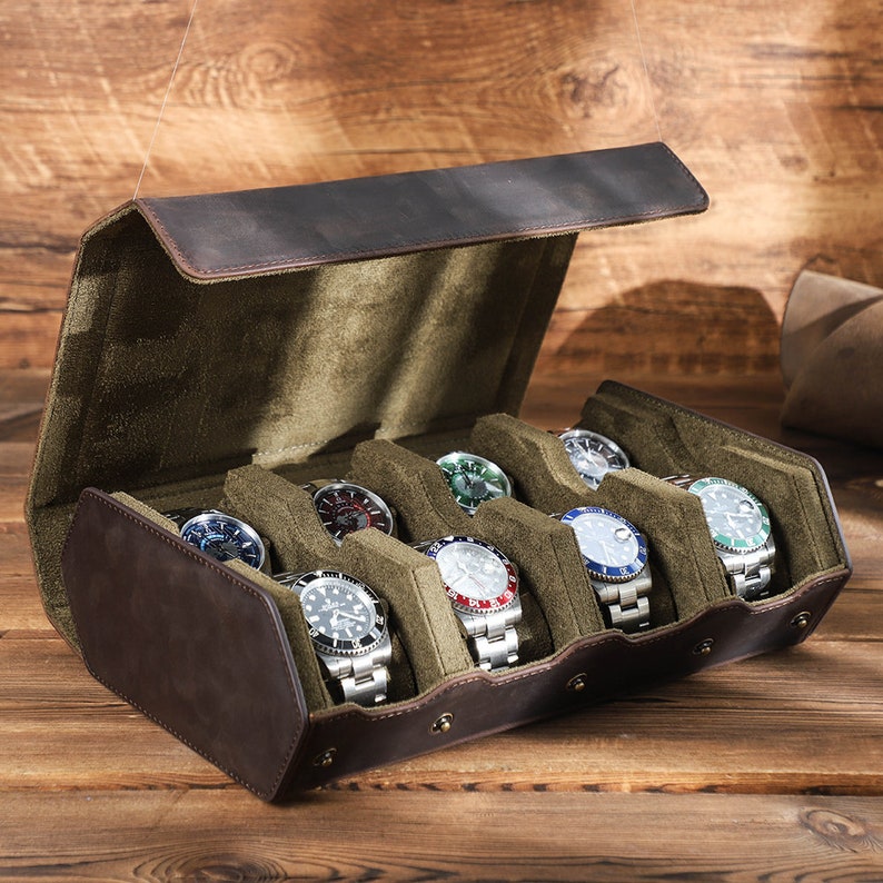 May include: A brown leather watch box with a green velvet interior. The box has six slots for watches and is open to reveal the watches inside. The watches are all different styles and colors.