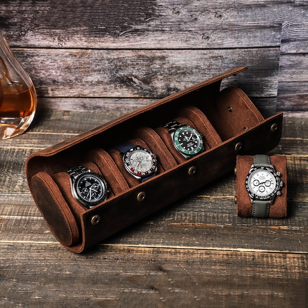May include: A brown leather watch roll with five slots for watches. The roll is open and four watches are visible. The watches have silver, black, and green faces. The watch roll is on a wooden surface.