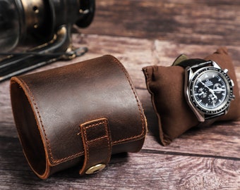 May include: A brown leather watch roll with a snap closure. The roll is open and a silver watch with a black face is resting on a brown cushion.