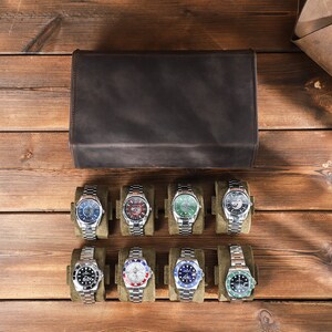 May include: Eight silver wristwatches with different colored faces and bands are displayed in a row on a brown wooden surface. The watches are arranged in two rows of four, with each watch in a separate compartment. The watches have a variety of colored faces, including blue, green, red, and white. The bands are all silver.