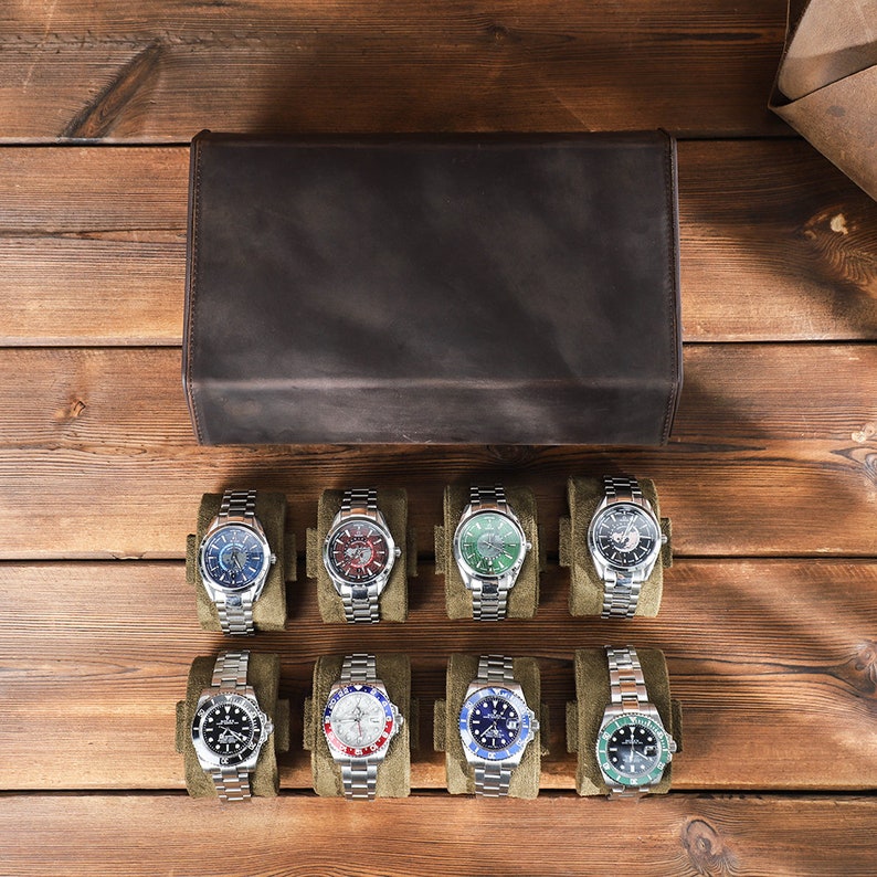 May include: Eight silver wristwatches with different colored faces and bands are displayed in a row on a brown wooden surface. The watches are arranged in two rows of four, with each watch in a separate compartment. The watches have a variety of colored faces, including blue, green, red, and white. The bands are all silver.