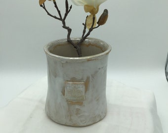 Può includere: Un vaso in ceramica bianca con una superficie testurizzata e un design decorativo. Il vaso è riempito con un singolo ramo di fiori di magnolia bianchi.