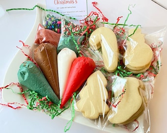 May include: A Christmas DIY cookie kit presented on a white platter. The kit contains festive-shaped sugar cookies, icing bags, and colourful sprinkles. The kit is decorated with red and green paper shreds and a card that says "Christmas's DIY cookie kit instructions."
