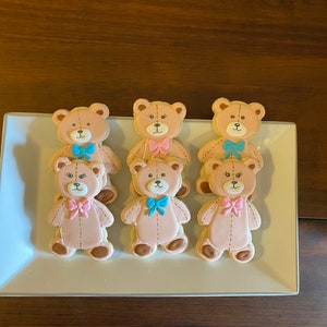 May include: Six decorated sugar cookies shaped like teddy bears. The cookies are arranged in two rows of three on a white rectangular plate. The teddy bears are all brown and have pink or blue bows.