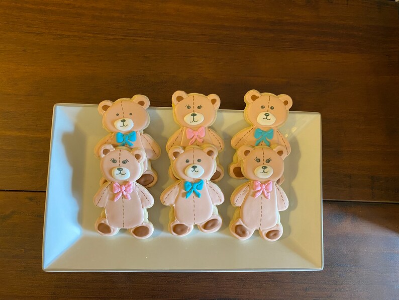 May include: Six decorated sugar cookies shaped like teddy bears. The cookies are arranged in two rows of three on a white rectangular plate. The teddy bears are all brown and have pink or blue bows.