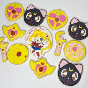 May include: A set of ten decorated sugar cookies featuring a Sailor Moon theme. The cookies are yellow, pink, and black with various designs including a crescent moon, stars, and a cartoon character. The text "Happy Birthday Aralin!" is written on one of the cookies.
