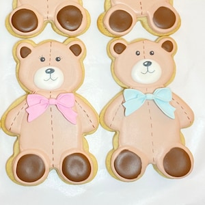 May include: Two decorated sugar cookies shaped like teddy bears. One bear is wearing a pink bow tie and the other is wearing a blue bow tie.