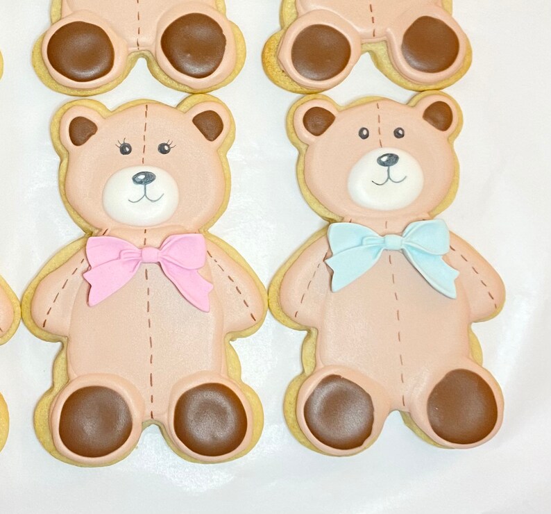 May include: Two decorated sugar cookies shaped like teddy bears. One bear is wearing a pink bow tie and the other is wearing a blue bow tie.