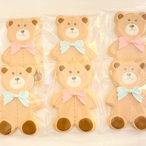 May include: Six decorated sugar cookies shaped like teddy bears. The cookies are brown with white faces and brown eyes. Each cookie has a bow tied around its neck. Three cookies have blue bows and three cookies have pink bows.