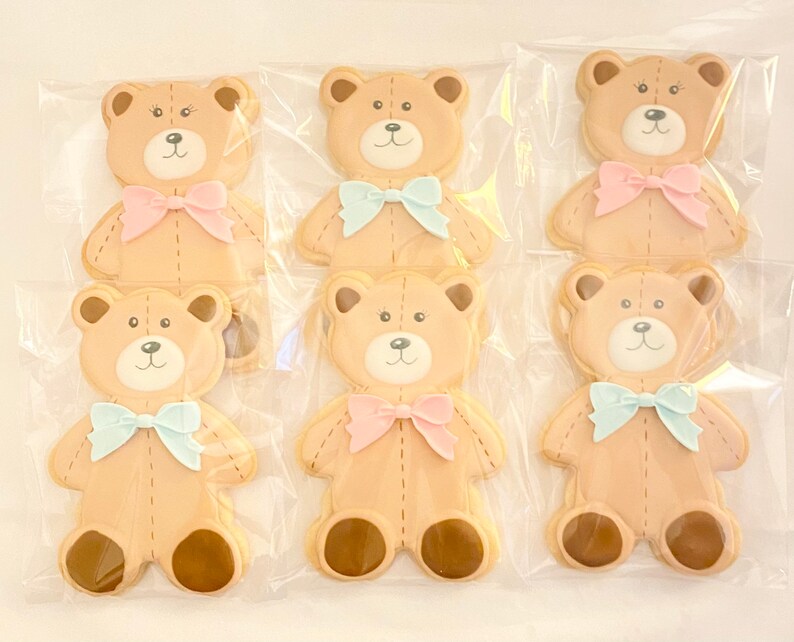 May include: Six decorated sugar cookies shaped like teddy bears. The cookies are brown with white faces and brown eyes. Each cookie has a bow tied around its neck. Three cookies have blue bows and three cookies have pink bows.