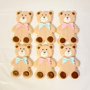 May include: Six decorated sugar cookies shaped like teddy bears. The cookies are brown with white faces and brown eyes. Each cookie has a bow tied around its neck. Three cookies have pink bows and three cookies have blue bows.